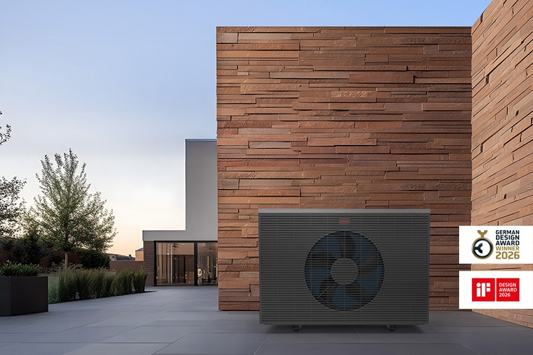 Modern exterior view with a large, dark-coloured AEROTOP SPK heat pump in front of a tall, textured brick wall.